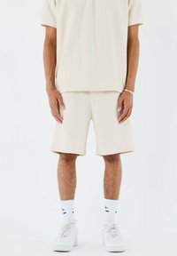 Camisa beige de manga corta y pantalones cortos a juego en tejido texturizado, con un ajuste relajado. Combinados con calcetines blancos y zapato deportivo.