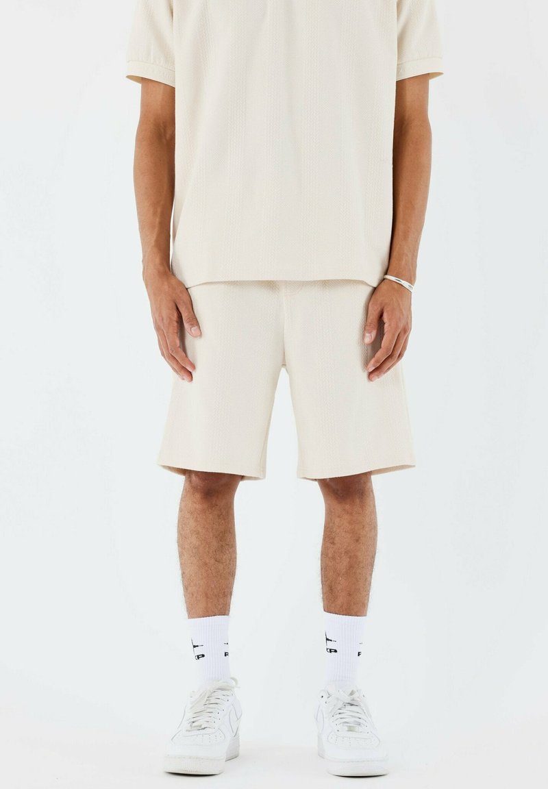 Camisa beige de manga corta y pantalones cortos a juego en tejido texturizado, con un ajuste relajado. Combinados con calcetines blancos y zapato deportivo.