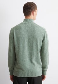 Grüner strukturierter Strickpullover mit hohem Kragen und geripptem Saum. Der Stoff hat subtile Einschlüsse und eine lockere Passform. Sichtbare Nähte an den Schultern.