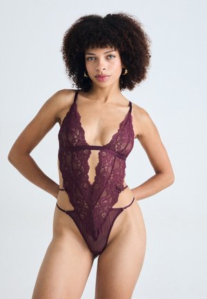 Donna con capelli ricci che indossa un bodysuit di lingerie in pizzo viola scuro con tagli, in piedi davanti a uno sfondo chiaro e uniforme.