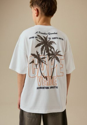 Dreng iført hvid T-shirt med brunt palmetrædesign og teksten "Cape Verde Adventure Awaits" på ryggen, stående mod beige baggrund.