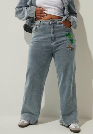 Persoon draagt lichtblauwe wijde jeans met palmboom- en luipaardborduursel, een wit cropped topje, een spijkerjack en witte sneakers.