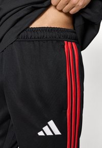 Μαύρο αθλητικό παντελόνι με κόκκινες λωρίδες στο πλάι, ελαστική ζώνη μέσης και λευκό λογότυπο. Κατασκευασμένο από απαλό ύφασμα με άνετη εφαρμογή.