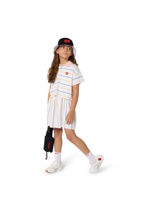 Robe polo rayée en blanc, jaune et rose, ornée d'un logo en forme de cœur. Associée à un bob noir et des baskets blanches.