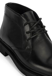 Bottines en cuir noir avec une texture lisse, un talon bas et un design classique à lacets. Elles présentent une pointe arrondie et de subtils accents en couture.