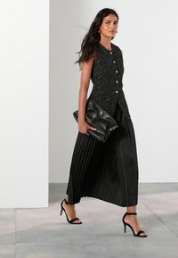 Czarna plisowana spódnica i dopasowany, bezrękawnik o fakturze z guzikami. Modelka trzyma czarną kopertówkę i ma na sobie czarne sandały na obcasie z paskami.