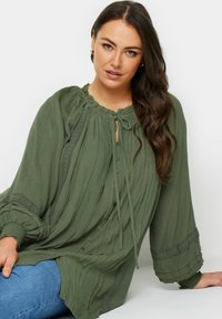 Grön blus med långa ärmar, med en rynkad halsringning, knäppning med knappar och spetsdetaljer; tillverkad av lätt, strukturerad tyg.