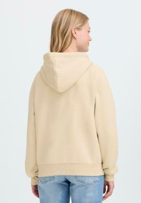Beige hoodie with a drawstring hood, ribbed cuffs, and hem; soft texture; worn with light blue jeans; shown from the back.
