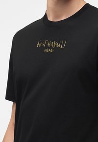 Μαύρο μπλουζάκι με μικρό χρυσό λογότυπο "Just Cavalli Milano" στο στήθος, που φοριέται από ένα άτομο, εμφανιζόμενο από τον λαιμό μέχρι το μέσο του κορμού.