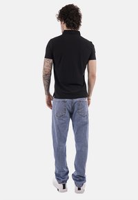 Homme aux cheveux bouclés, bras gauche tatoué, portant un polo noir, un jean bleu et des baskets blanches, debout de dos sur un fond blanc.