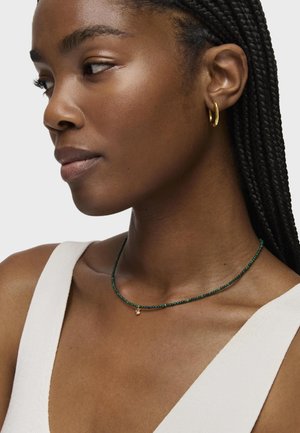 Collana di perle verdi con un ciondolo d'oro, indossata sulla pelle nuda. Il design presenta piccole perline rotonde e una delicata catena.