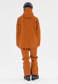 Wasserdichte orange Skijacke und -hose, ausgestattet mit einer Kapuze, langen Ärmeln und verstellbaren Bündchen. Abgerundet mit schwarzen Skischuhen.