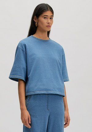 Vrouw met lang donker haar, gekleed in een loszittend blauw denim topje met korte mouwen en bijpassende broek, staand tegen een effen lichte achtergrond.