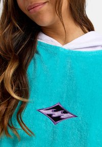 Tyrkysový froté pončo s bielou kapucňou a čiernym diamantovým logom s vyšitými fialovými vlnami na hrudi.