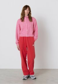 Žena stojí proti bielej stene, oblečená v ružovom kardigane na gombíky, červených teplákoch s logom a šedých teniskách, pózuje s rukami v vreckách.