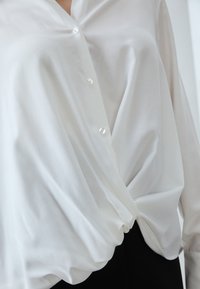 Närbild av en vit, långärmad blus med knappar och en tvinnad knutdetalj i midjan, som bärs över svarta byxor.