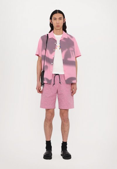 Korte mouwen roze en grijze overhemd met knopen, witte graphic t-shirt, roze gebloemde shorts, zwarte schoenen en een zwarte schoudertas.