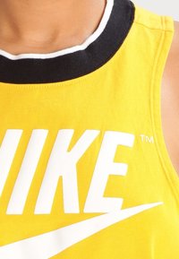 Žluté tank top Nike s černobílým žebrovým límcem. Na přední straně se nachází velké bílé logo "NIKE" a design swoosh.
