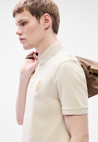 Μπεζ πόλο πουκάμισο με γιακά και κοντά μανίκια, με κεντημένη διακόσμηση. Μια καφέ τσάντα είναι περασμένη πάνω από τον έναν ώμο.