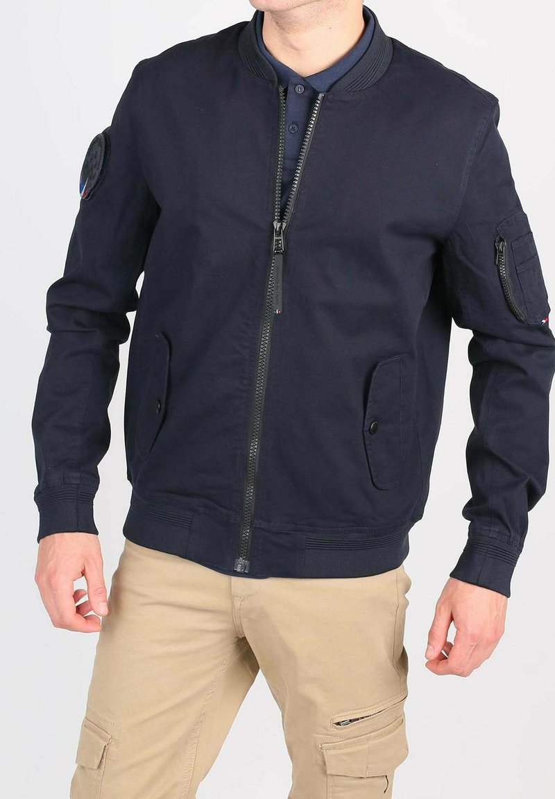 Homme portant une veste bomber bleue marine avec des poches zippées et à rabat, un pantalon cargo beige avec des poches zippées et à rabat, sans tête visible.