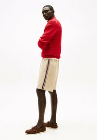 Maglione rosso lavorato a maglia, pantaloni corti beige con strisce laterali blu navy, rosse e bianche, e scarpe da barca marroni. Modello posato di lato contro uno sfondo chiaro.