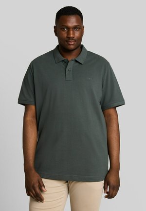Mann trägt dunkelgrünes kurzärmeliges Poloshirt und beige Hose, steht vor einem schlichten hellgrauen Hintergrund.