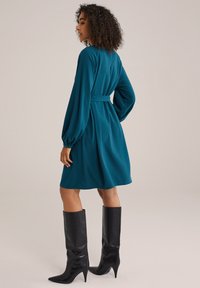 Teal långärmad klänning med insnörd midja, mjukt tyg och ballongärmar, i kombination med knähöga svarta spetsiga stövlar.