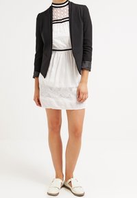 Blazer negro ajustado sobre un vestido blanco de encaje con escote redondo, que presenta detalles en negro. La modelo lleva zapatos blancos tipo slip-on.