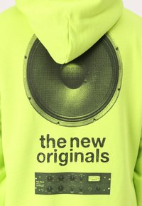 Neon grüne Kapuzenjacke mit einem großen schwarzen Lautsprecher-Graphik und dem Text "die neuen Originals" über einer Grafik eines Kontrollpanels.