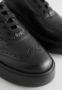 Musta nahkakengät, joissa on brogue-muotoilu koristeellisilla rei'illä ja teksturoidulla pinnalla, ja paksu kumipohja.