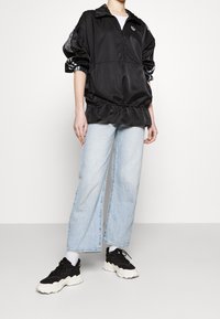 Person klädd i en svart halvdragkedjajacka med mönstrade ärmstripes, ljusblå vida jeans, vita strumpor och svarta tjocka sneakers som står på grått golv.