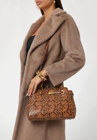 Bolso con estampado de serpiente marrón con detalles en hardware dorado, con forma estructurada y correa de hombro removible, sostenido por una persona con un abrigo beige.