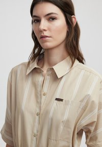 Camicia beige con bottoni, con strisce verticali bianche, taschino sul petto e una piccola placca metallica con logo. Tessuto morbido e maniche corte.