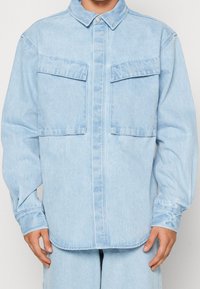 Light blue denim shirt with a button-up front, two chest pockets, long sleeves, and a relaxed fit. Features a pointed collar and cuffed sleeves.