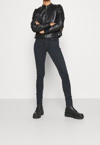 Schwarze Lederjacke mit Reißverschluss und Schulterdetails, kombiniert mit dunklen Slim-Fit-Jeans und schwarzen Plate Stiefeln, die einen monochromen Look erzeugen.