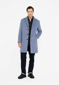 Mann in einem mittel-langen blauen strukturierten Mantel, schwarzem Hemd, dunklen Jeans mit Umschlägen und schwarzen Schuhen vor einem weißen Hintergrund stehend.