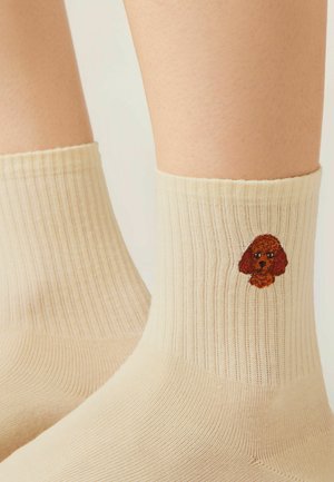 Beige geribbelde sokken gedragen op de voeten, elk met een kleine geborduurde bruine hondenkop aan de buitenkant nabij de enkel.