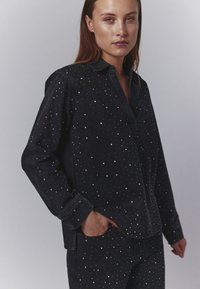 Cămașă din denim negru cu guler cu nasturi și embellismente din nasturi argintii. Mâneci lungi cu mansete, croi lejer și buzunare laterale vizibile.