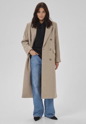 Frau mit dunklem Haar, die einen beigen langen Mantel über einem schwarzen Hoodie, blauen Jeans und schwarzen spitzen Schuhen trägt und vor einem schlichten Hintergrund steht.