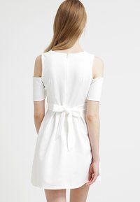 Vestido blanco con hombros descubiertos, cuerpo ajustado y falda acampanada. Presenta un detalle de lazo en la espalda y un cierre de cremallera discreto.