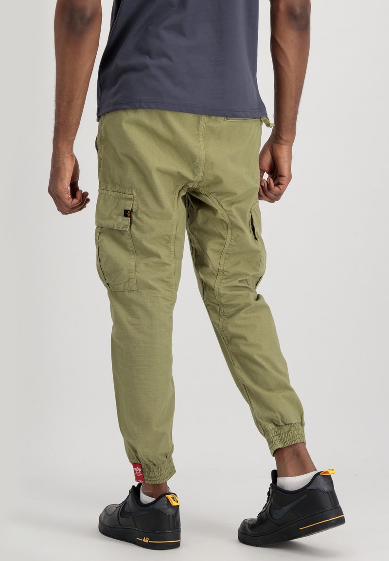 Zielone spodnie cargo mają elastyczne mankiety i kieszenie po bokach, w zestawieniu z czarnymi trampkami. Tkanina ma lekką, teksturowaną powierzchnię.