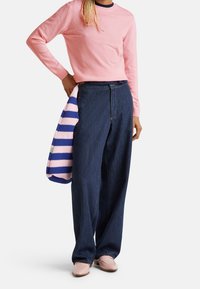 Person som bär en rosa tröja, mörkblå vida jeans och rosa skor, medan han/hon håller en stickad väska i rosa och blå ränder.