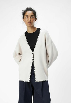 Lichtbeige cardigan met zwarte zoom, met een V-hals, open voorkant en een iets oversized pasvorm, gecombineerd met een zwarte top en losse donkere broek.