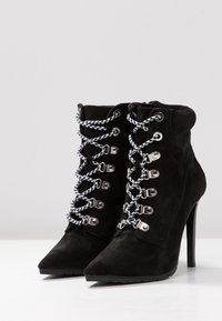 Black suede ankle boots with pointed toes and high heels. Featuring white and black patterned laces and silver hardware accents.