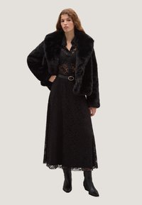 Giacca in finta pelliccia nera con ampi risvolti, indossata sopra un vestito in pizzo nero con intricati motivi floreali e una cintura nera elegante. Stivali neri con tacco.