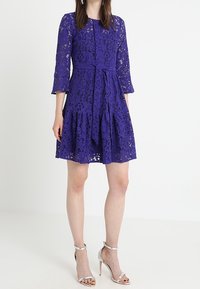 Fialové krajkové šaty s tříčtvrtečními rukávy, přiléhavým pasem, rozšířenou sukní a detailem s vázaným páskem. Hodí se k nim stříbrné sandály na vysokém podpatku.