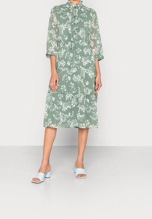 Person trägt ein grünes Midi-Kleid mit weißem Blumenmuster, durchgeknöpft, mit Bindegürtel, dreiviertel Ärmeln und hellblauen Blockabsatz-Sandalen mit offenem Zehenbereich.