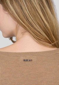 Pull en tricot marron avec un large col et une étiquette de marque métallique à l'arrière. Texture douce et coupe décontractée.