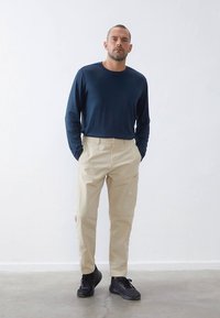 Mornarsko modra majica z dolgimi rokavi, bež hlače z žepi, teksturirana tkanina, obuta v črne superge, stoji ob nevtralni podlagi.