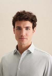 Lichtgrijze button-up shirt van een gestructureerde stof, met een puntige kraag en witte knopen. Het model heeft kort, krullend haar en blauwe ogen.